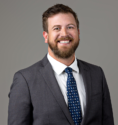 Mack wilding, pictured in a professional headshot, a white man with short brown hair, a brown beard, smiling, in a gray suit with a blue tie and a gray background.