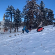 A photo of three teens and pre-teens running up a snow-covered hillside with sleds. There is a stand of ponderosa pine trees, the Great Stupa of Dharmakaya, and clear blue skies in the background.