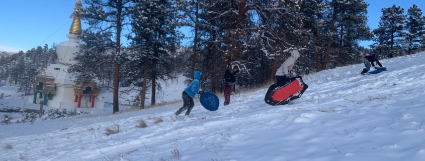 A photo of three teens and pre-teens running up a snow-covered hillside with sleds. There is a stand of ponderosa pine trees, the Great Stupa of Dharmakaya, and clear blue skies in the background.